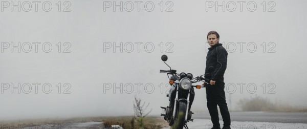 A young man stands by a motorcycle on a foggy road, looking ahead