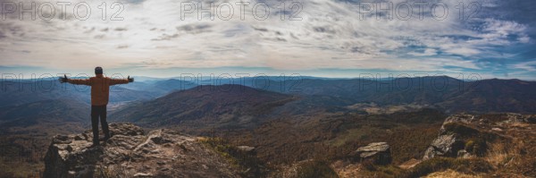 Panoramic view of mountain valley. Young man standing with hands widespread trying to embrace whole world. Man is a small part of astonishind mountain landscape with cloudy sky