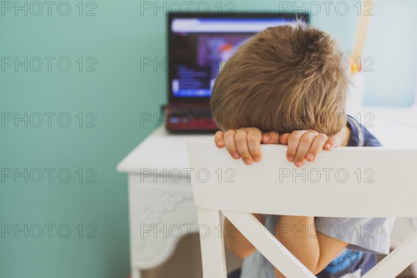 The sad little boy sits in front of laptop and doesn't want to look lesson. Concept of boring learning at home