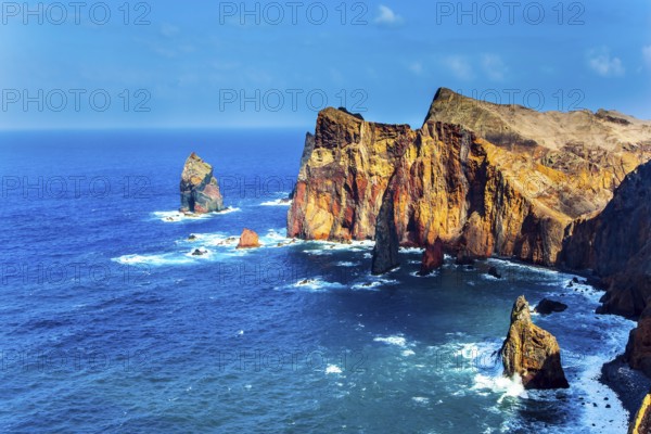 Island in the Atlantic. The coast of the island of Madeira - red rocks, azure surf. The blinding midday sun illuminates the rocky shore. The concept of active, ecological and photo tourism