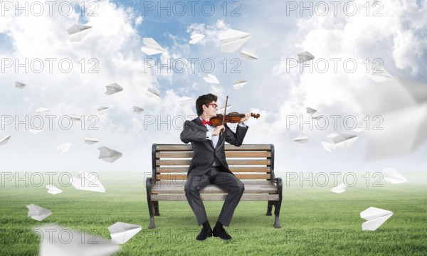 Young businessman plays the violin sitting on a bench, around flying paper planes. Creativity in business