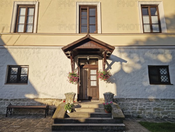 Rustic old building facade with porch and a charming wooden door, framed by two side windows and a small overhang. Stone steps lead up to the entrance, adorned with flower pots in bloom