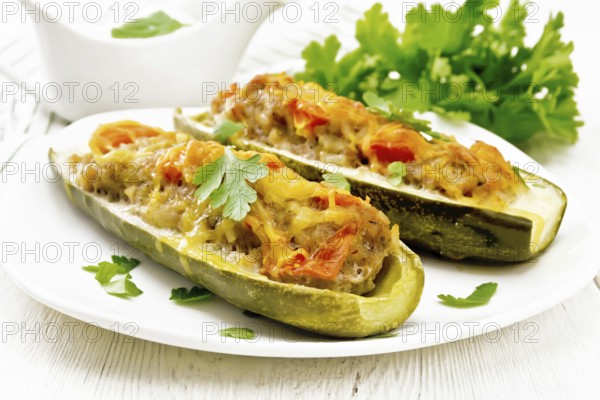 Cucumbers stuffed with minced meat, tomatoes, pepper with cheese and parsley in a plate, cream and greens sauce in a gravy boat, towel on white wooden board background