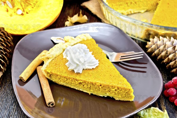 A piece of pumpkin pie with whipped cream in a brown plate with a fork and cinnamon, pumpkin, cones, berries of viburnum, maple leaf on the background of dark wood planks