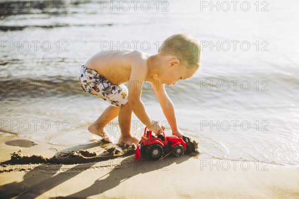 Caucasian child boy plays toy red tractor, excavator on sandy beach by the river in shorts at sunset day
