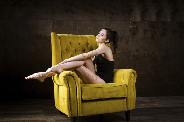 A young beautiful, sexy Caucasian woman with thin figure and long bare legs, barefoot posing reclining on yellow armchair in the interior against dark wooden wall. Dressed in a black classic swimsuit