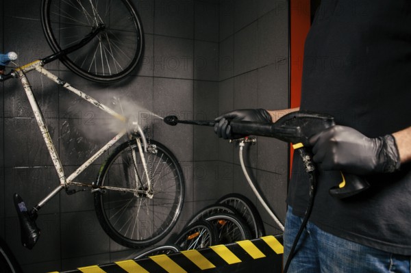 Services professional washing of bicycle in the workshop. Close-up of hand Young Caucasian stylish man doing bicycle cleaning using an automatic electric water pump. Sprays scatter from the pressure