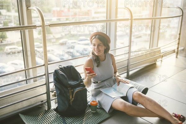 Theme of tourism and travel of young student. Beautiful young caucasian girl in dress and hat sits on floor tourist rug inside terminal airport terminal. Waiting room delayed flight, delay departure