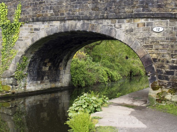 An old stone bridge crossing the rochdale canal at mytholmroyd west yorkshire with a waterside pathway surrounded by trees and bushes