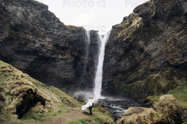 The groom circles the bride in his arms near the waterfall, it is snowing. Destination Iceland wedding, near Kvernufoss waterfall
