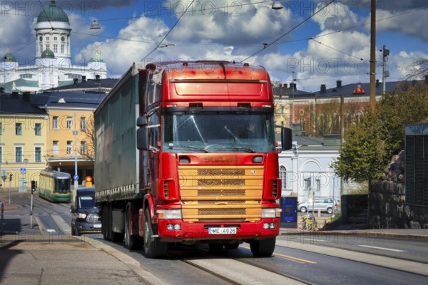 Red and gold 4-series Scania truck pulling semi trailer to Katajanokka Harbour. On the background city of Helsinki, Finland. April 29, 2020