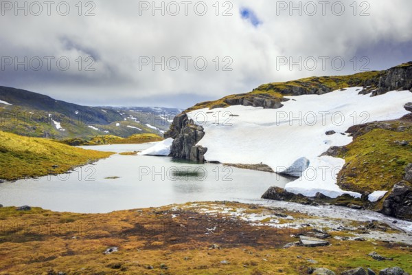 The cold water of the lake is covered with ice floes. Gorgeous Snow Road in Norway. Cold July in Northern Europe. The cold snowfields and sharp stone placers