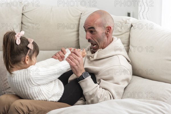 Bald father playing with his daughter on the sofa, making funny faces and holding her feet