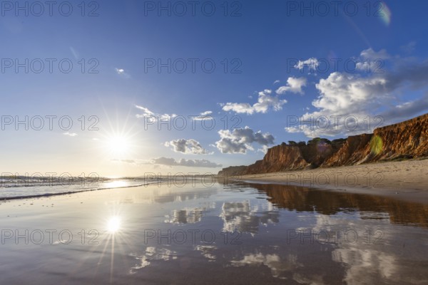 The golden sandy beach of Praia da Falésia, crystal clear sea water at low tide with the reflection of the sky, the setting sun and the characteristic orange-coloured cliffs, Algarve, Portugal