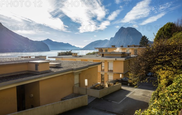 Amazing view of 4-story apartment buildings on the shores of Lake Lugano and a park on the coast with palm trees and a brilliant Lake Lugano and white pebbles on the roof of houses and Mount San Salvatore