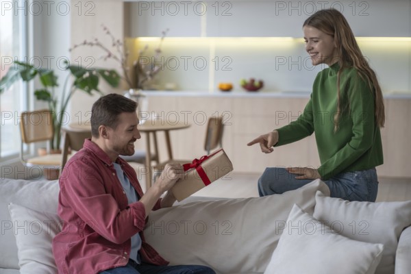 Happy couple celebration. Loving pleased woman gives gift with red tape to smiling husband. Satisfied man holding present box from girlfriend on birthday feeling interest and anticipation at home