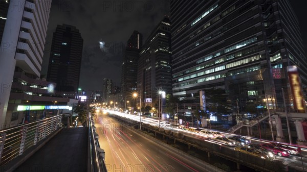 Timelapse shot of night Bangkok street. Intense transport traffic on the road with highrise buildings alongside. The moon and sailing clouds in dark sky