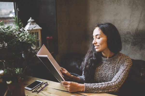 Young business girl studying the menu in restaurant decorated with Christmas decor.sits near the window on cloudy winter day at wooden table.Dressed in warm grey jumper.On the table, phone and glasses