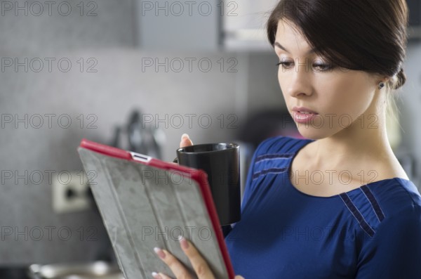 Portrait of young attractive caucasian brunette housewife at kitchen. Morning with cup of coffee and tablet pc