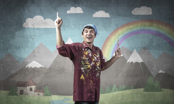 Young male artist with paintbrush. Happy painter in shirt and bandana standing on background colorful picture on wall. Summer landscape with mountains and rainbow. Creative hobby and profession