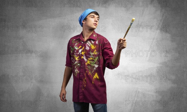 Young emotional artist gesturing with paintbrush. Caucasian painter in dirty shirt and bandana standing on gray wall background. Creative hobby and artistic occupation concept with copy space