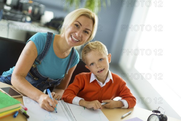 Young blonde caucasian kid in orange shirt and his mother sit at the table with colorful folders and alarm clock and draw together, look at camera Blurred office on background