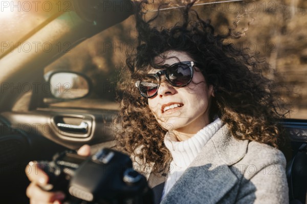 A relaxed woman with sunglasses holding a camera in a sunny convertible ride