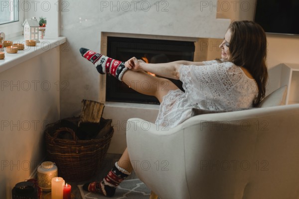 The young girl is sitting in a cozy room by the fireplace. The girl is sitting in an easy chair and dresses warm christmas socks