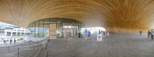 Calgary, Alberta, Canada. Aug 10, 2024. The Calgary Library entrance showing a spacious, modern area with a curved wooden ceiling, expansive glass walls, and a few people gathering outside