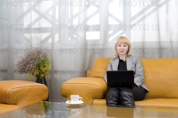 Young blonde woman sits on leather sofa with laptop, in front of glass table
