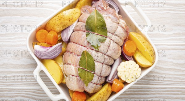 Raw wrapped rolled sliced pork in white casserole dish with potatoes, vegetables and herbs on rustic white wooden background top view. Pork roll with vegetables ready to be prepared, food photography, food photography
