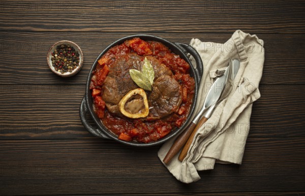 Traditional Italian dish Ossobuco all Milanese made with cut veal shank meat with vegetable tomato sauce served in black casserole pan top view on rustic brown wooden background, food photography, food photography