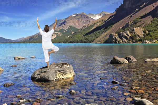 Yoga on the lake. Mature woman in white performs asana tree, standing on a rock