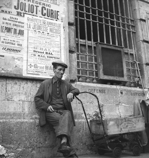 France in the 1950s. A man in the street is sitting having a cigarette. He leans on a primitive cart, on the wall behind him billboards are visible for previous and updoming events. Cannes France 1950 Kristoffersson ref AZ60-11
