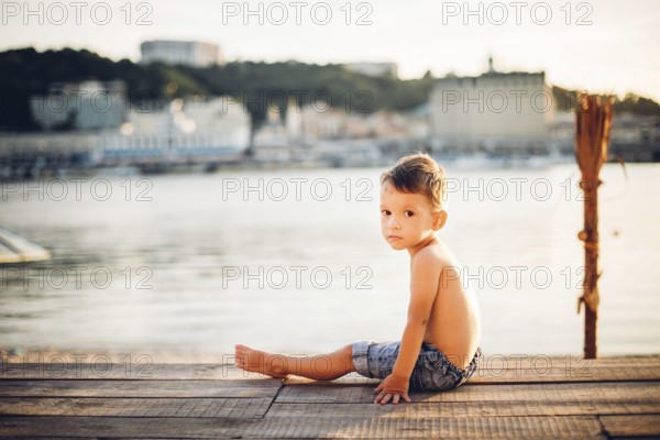 The theme is a child and summer beach holiday. A small Caucasian boy sits sideways on a wooden pier and looks at the camera on a sandy beach and a pond, a river. With bare legs in blue denim shorts