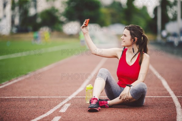 Young woman athlete at the stadium sporting lifestyle, sitting on the track, taking selfie photo on a smartphone, smiling joyful in her ears earpiece, near a bottle flask with water