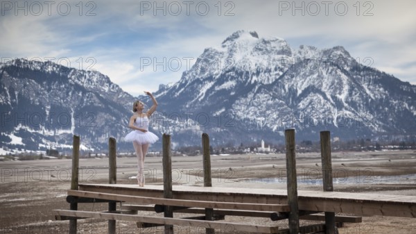 Ballerina dances elegantly on a wooden footbridge in front of snow-covered mountains, in the background the Säuling, Forggensee, Füssen, Allgäu, Bavaria, Germany