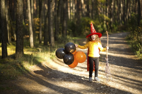 A girl in a witch costume and hat on a broom with orange and black balloons is playing in the autumn forest, going to a Halloween party