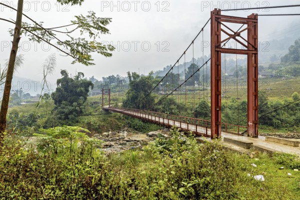 A red suspension bridge stretches across a green valley under an overcast sky, A bridge over a river in the countryside near Sa Pa in Vietnam