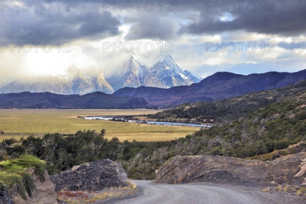 The road between turned yellow hills and river goes to snow-covered black rocks. Patagonia. Vertiginous landscape in the Chilean Andes