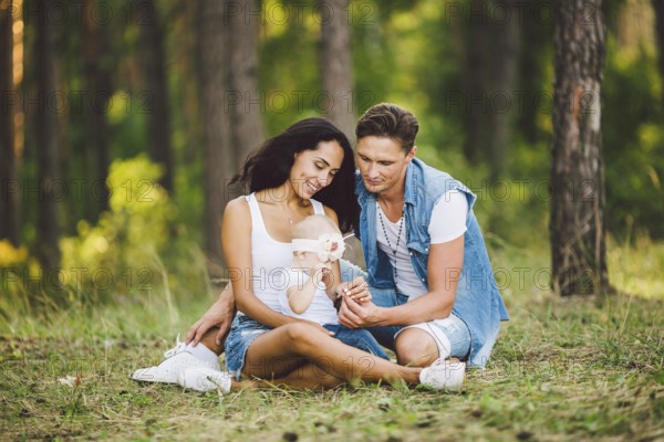 The theme family outdoor recreation in forest park area. Young beautiful stylish caucasian family with small child daughter one year old sit on the graß in the coniferous forest in summer at sunset