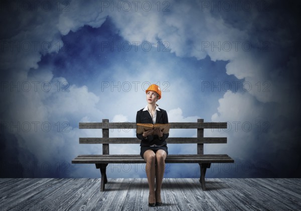 Beautiful woman architect sitting on wooden bench. Young technician specialist in orange safety helmet holding open book in hands on cloudy night sky background. Engineering and development company