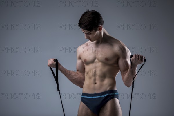 Muscular fit young male antique perfect muscles six packs of abs and bare chest. Bodybuilder model trains with a stretching elastic against a dark background in the studio. Workout training gym