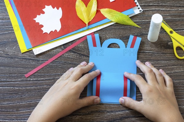 Instruction step 6. Greeting card paper backpack on a wooden table. Handmade. Project of children's creativity, handicrafts, crafts for kids