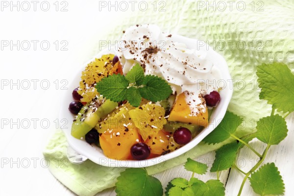 Fruit salad with orange, kiwi, cranberries and baked pumpkin, whipped cream, sprinkled with chocolate and coconut with mint in a bowl on a napkin on white wooden board background