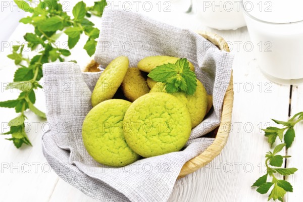 Green mint cookies on napkin in wicker basket, milk in a glass and a jug on white wooden board background