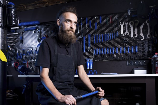 Portrait of small business owner of young man with beard. Guy bicycle mechanic workshop worker sitting with tool in his hand in a working black clothes in an apron in the background of a bicycle shop