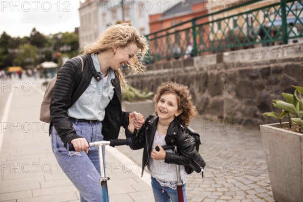 The young woman with a little girl are standing with scooters in the city, look each other