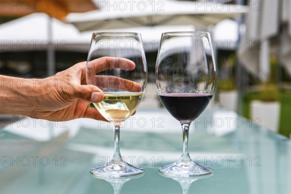 Man hand on white wine glass, pair of white and red wines glasses close up selective focus, restaurant patio outdoor table background