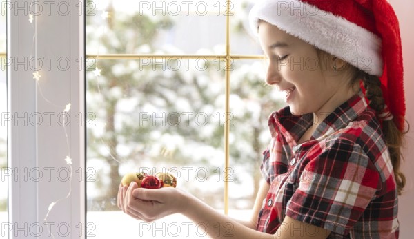 Girl in a Santa hat sits on the windowsill of a house near the Christmas tree and holding glass balls in his hands. Child is having fun, believes in a miracle and waiting for Christmas and New year